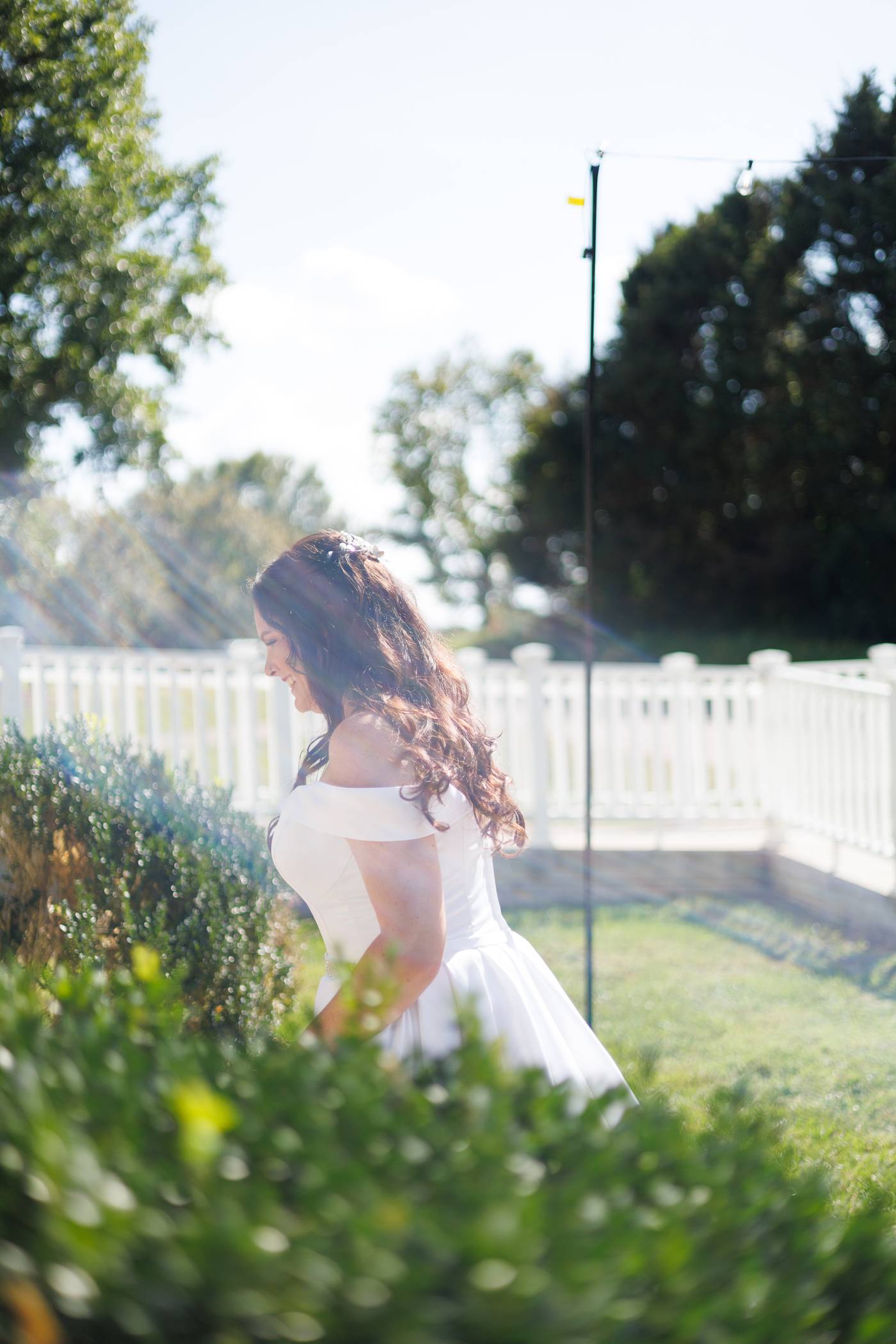 Bride in white dress smiles outdoors near a white fence and green bushes