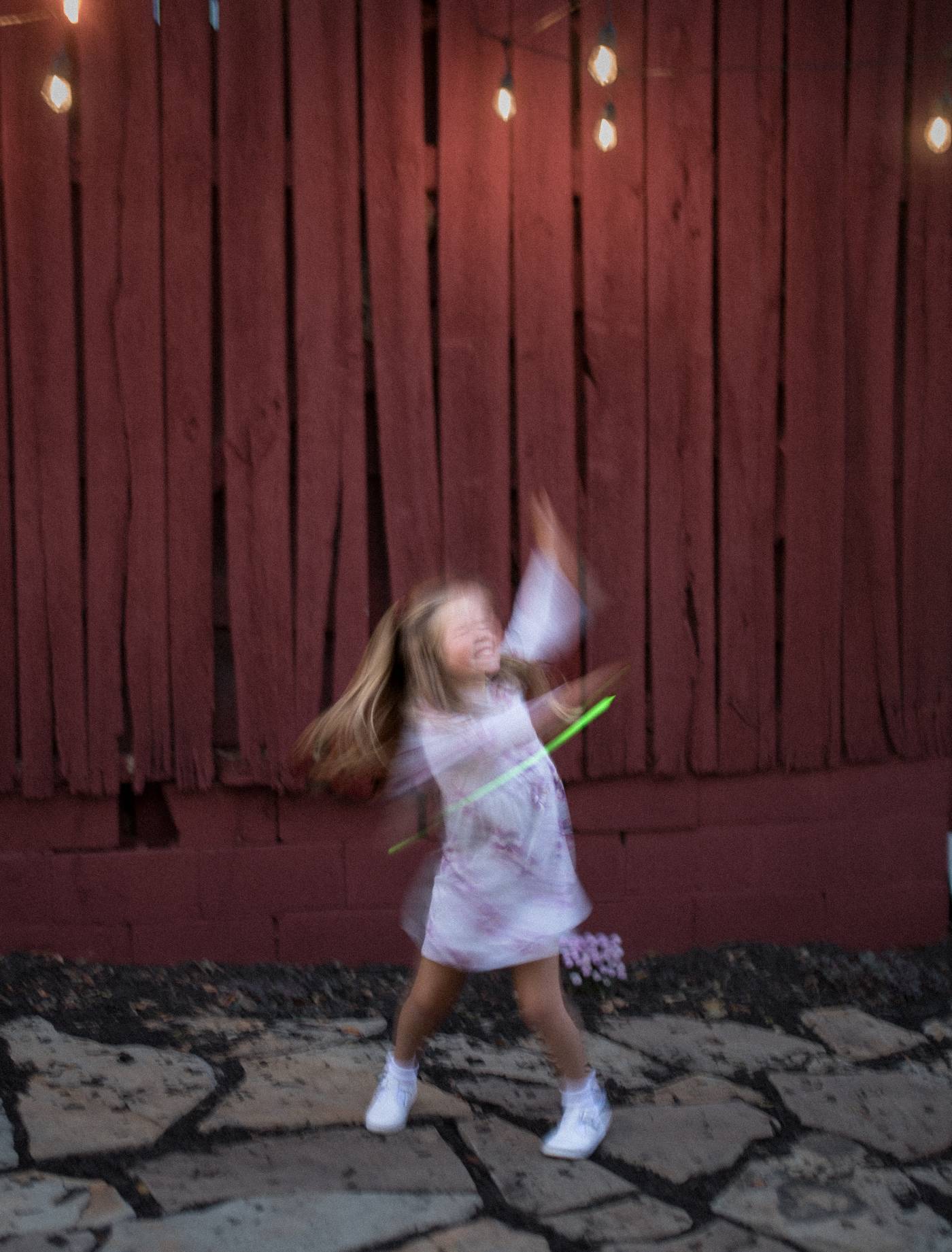 Joyful child twirls with glow sticks in front of a rustic wooden wall and string lights