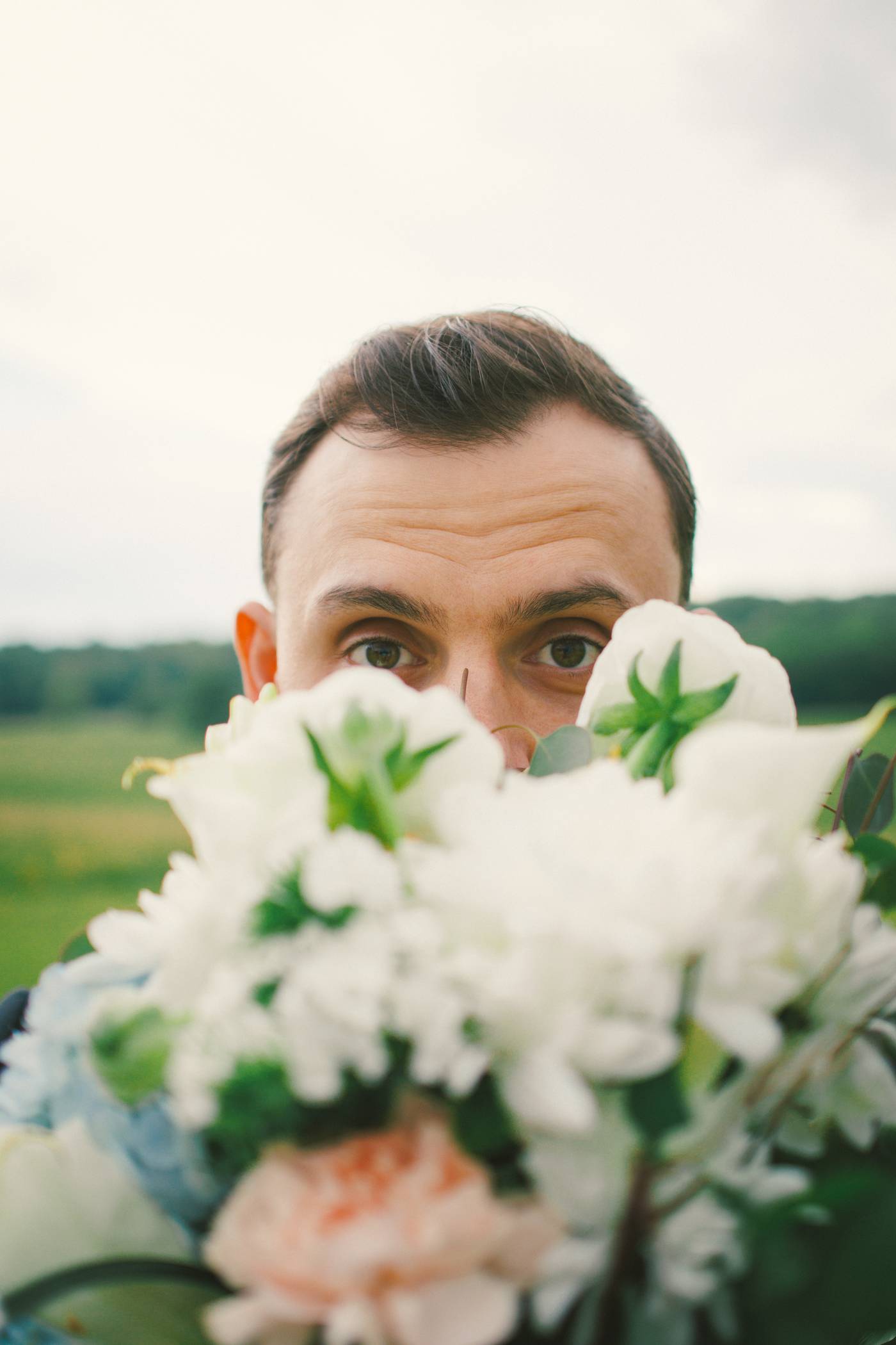 Groom peeking over a bouquet of white flowers outdoors on a cloudy day