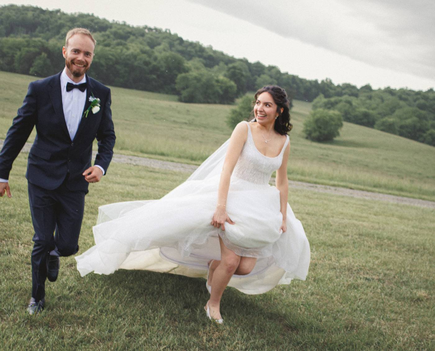 Smiling bride and groom run across a grassy hill on their wedding day