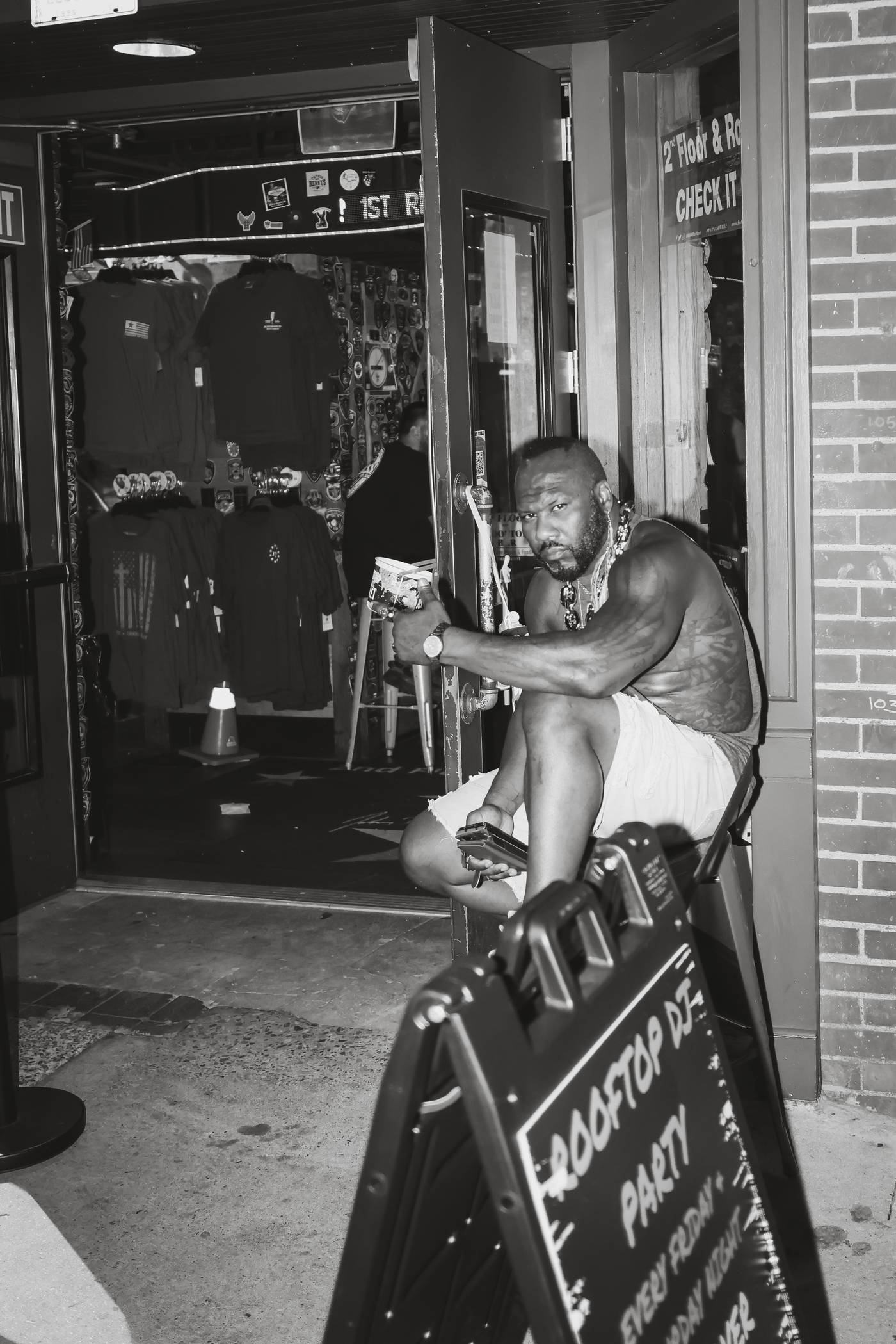 Man with tattoos and jewelry sits on a chair outside a store at night