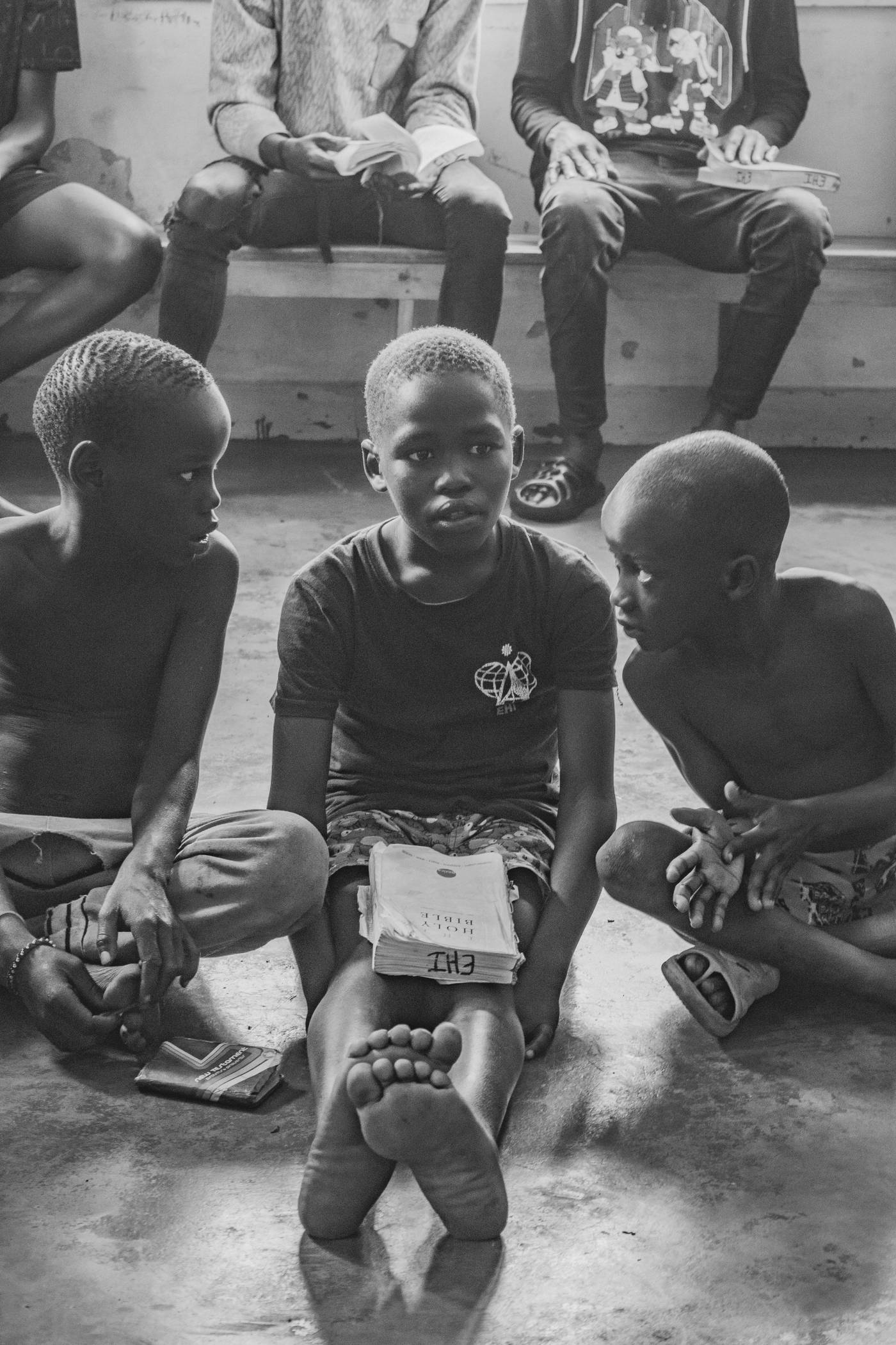Three boys sit on the floor, one holding a Bible, in a candid street scene