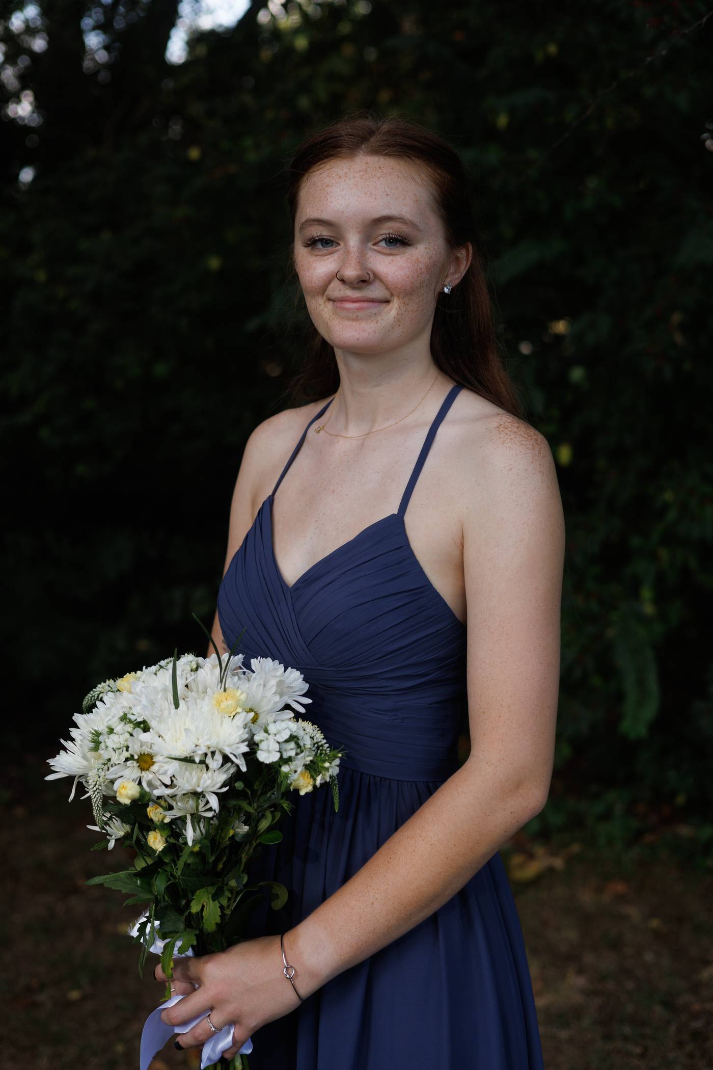 Bridesmaid in a navy dress holding a white daisy bouquet against dark greenery
