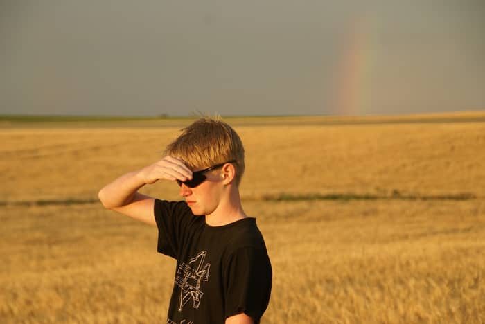 Teenager shields eyes in golden wheat field with faint rainbow overhead