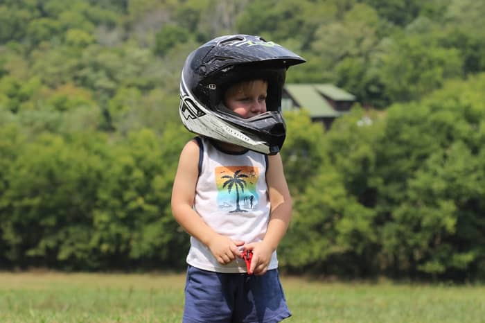 Young boy in motocross helmet stands in a grassy field with trees behind