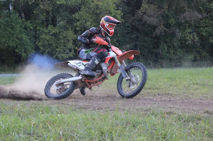 Dirt bike rider kicks up dust and smoke on a grassy track surrounded by trees