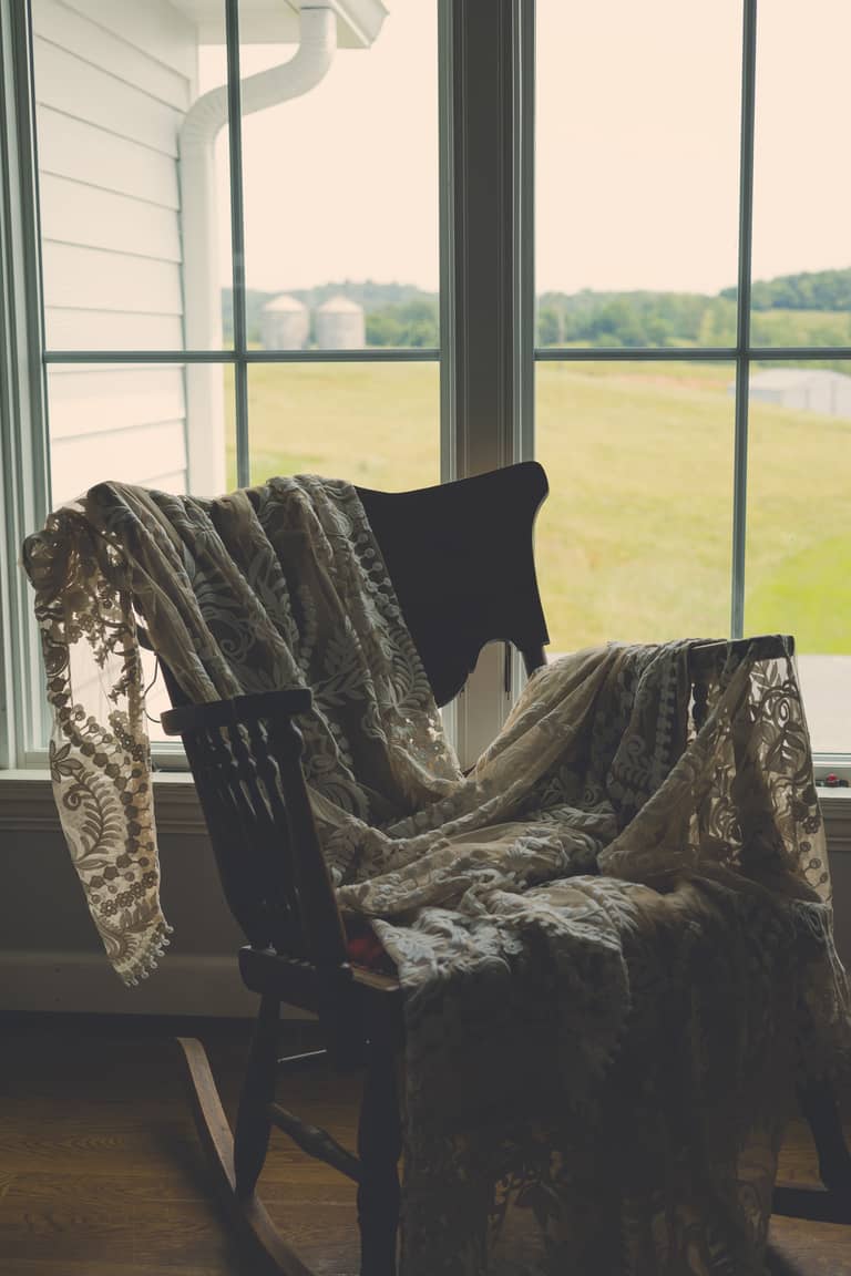 Lace wedding dress draped over a rocking chair by a window with a rural view