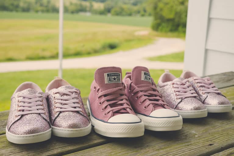 Several pairs of pink Converse sneakers sit on a wooden porch with a green field background
