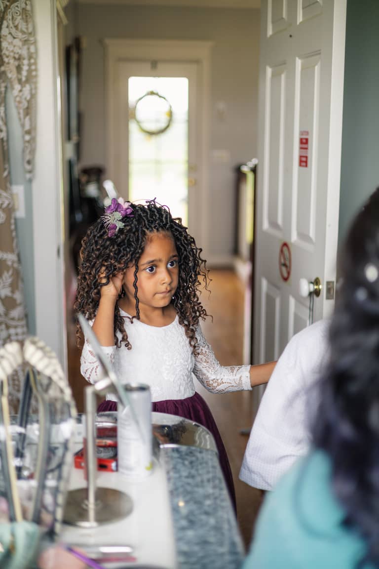 Flower girl with curly hair and purple flower accessory adjusts her earring before wedding