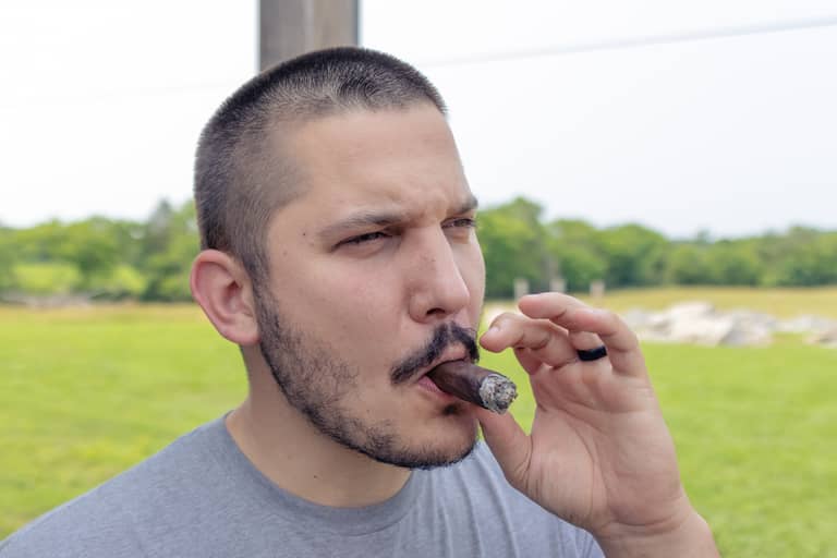 Groom enjoys a cigar outdoors with a blurred grassy background during a wedding shoot