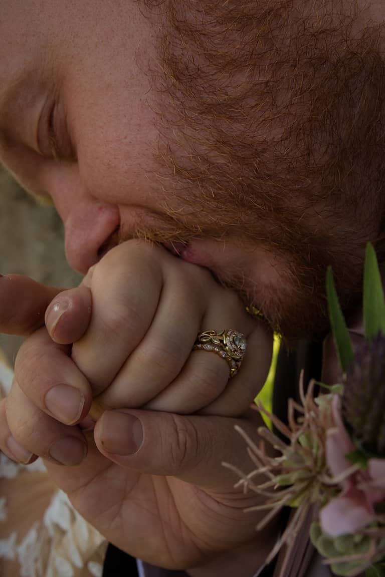 Groom kisses bride's hand, showing wedding rings and bouquet, intimate wedding moment