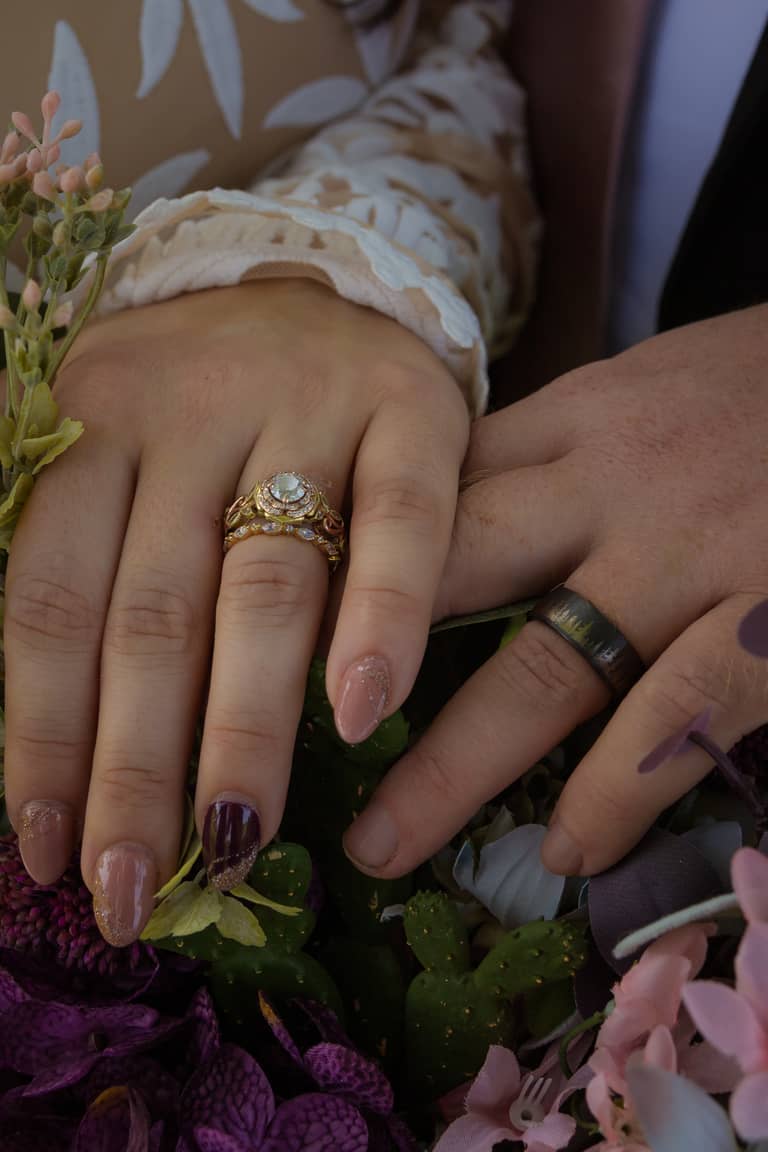 Wedding rings rest on hands amidst a bouquet of purple and pink flowers