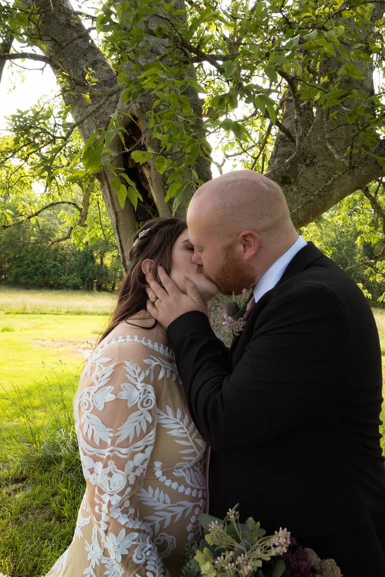 Bride and groom share a tender kiss under a large tree during their outdoor wedding ceremony