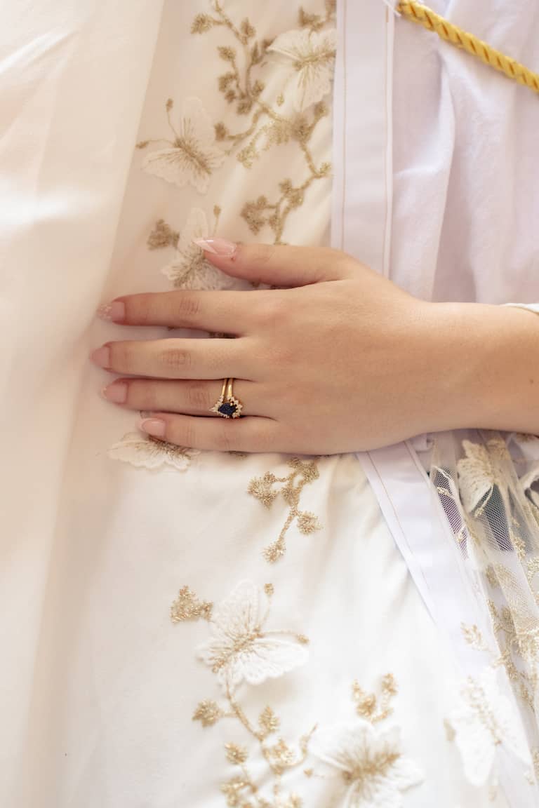 Bride's hand with a sapphire engagement ring rests on a white dress with gold embroidery