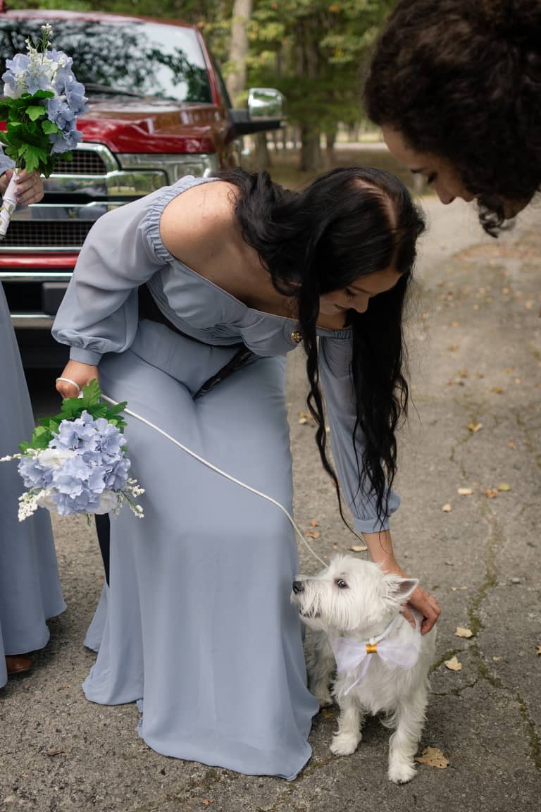 A bridesmaid pets a white dog with a bow during a wedding ceremony outdoors