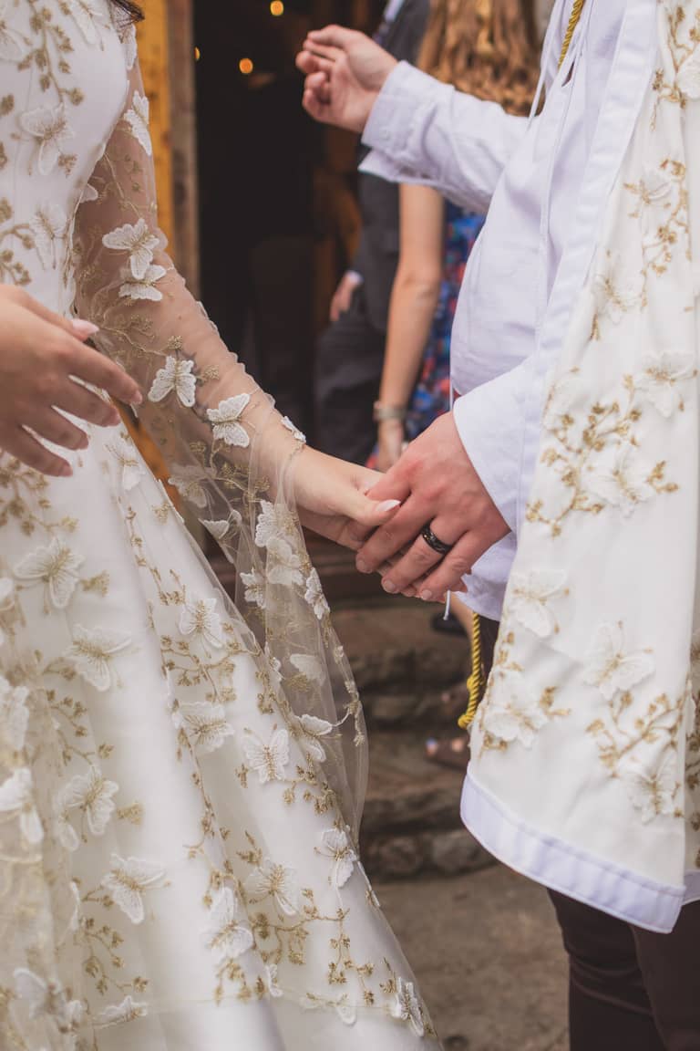 Bride and groom hold hands, wearing ornate white wedding attire outdoors