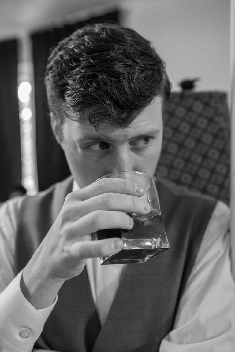 Groom in a vest sips a drink indoors, reflecting a moment of calm before the wedding