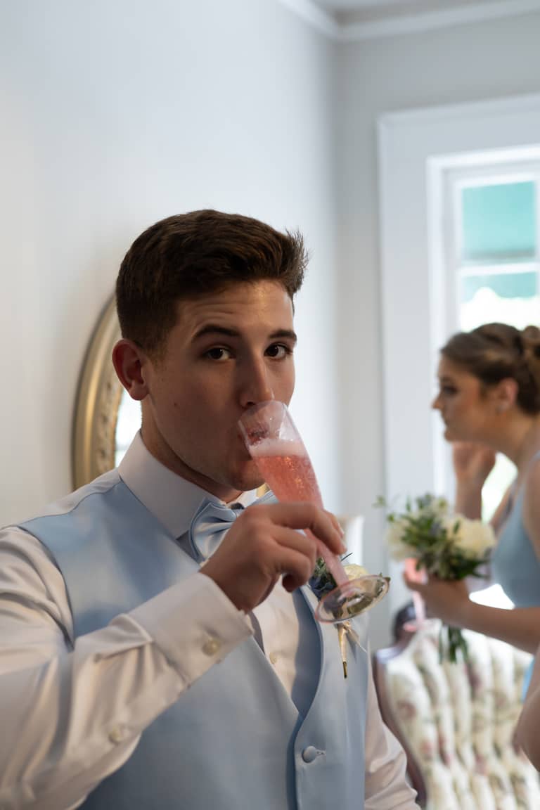 Groom in light blue vest drinks champagne, bridesmaid holds bouquet in background