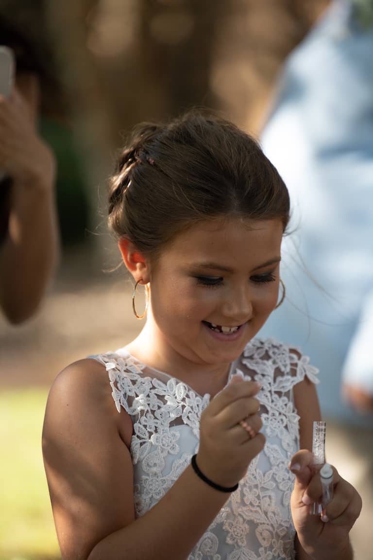 Flower girl in white lace dress smiles while holding small vials at an outdoor wedding