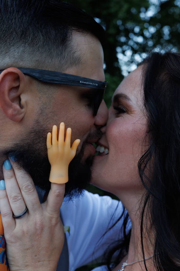 A bride and groom share a playful kiss outdoors with a novelty hand on his finger