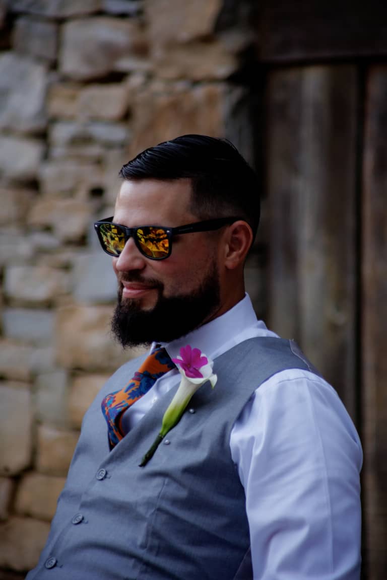 Groom in sunglasses and vest smiles outdoors at a wedding