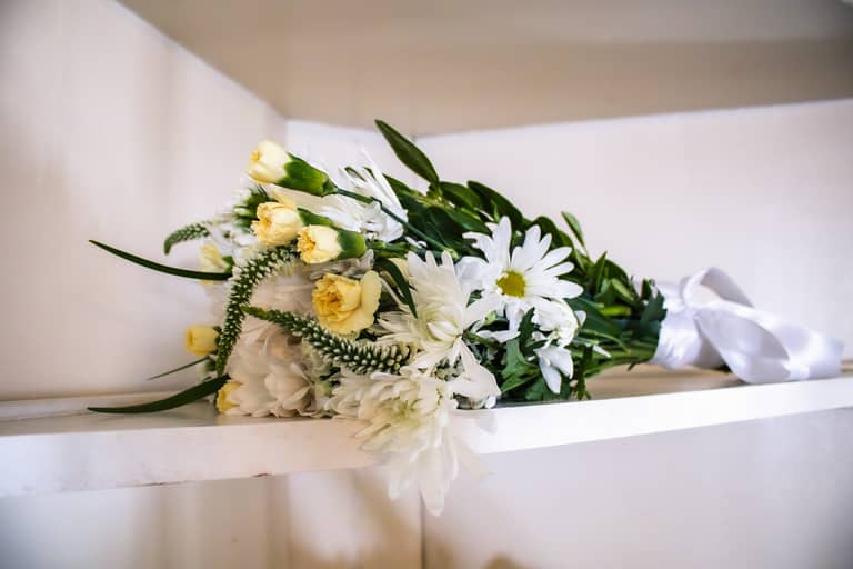 A wedding bouquet of white and yellow flowers rests on a shelf with a white ribbon