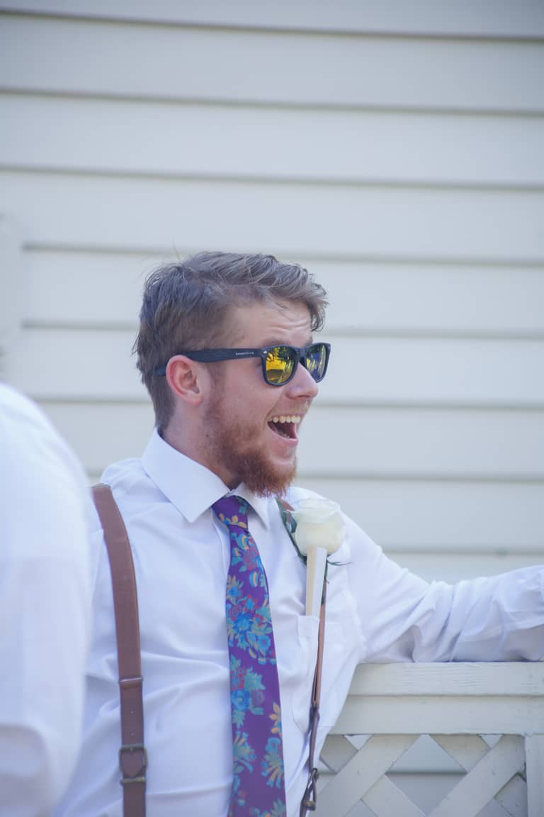 Groom in sunglasses and floral tie laughs outdoors during wedding celebration