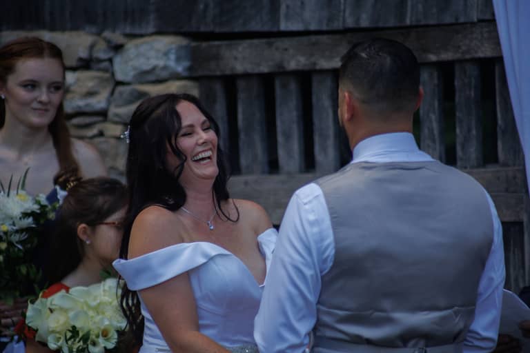 Bride laughs joyfully during a rustic outdoor wedding ceremony with guests in the background