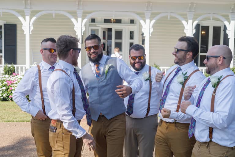 Groomsmen in suspenders and colorful ties laugh outside a white house during a wedding celebration
