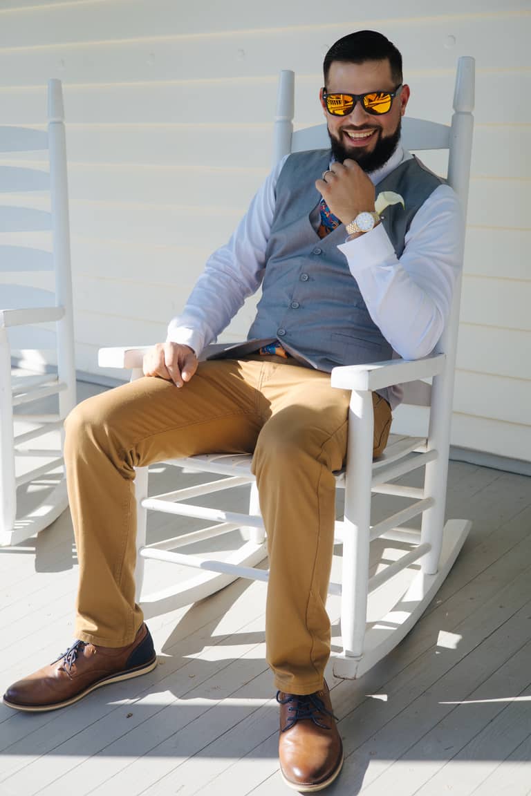 Groom in sunglasses smiles while sitting on a white rocking chair on a porch