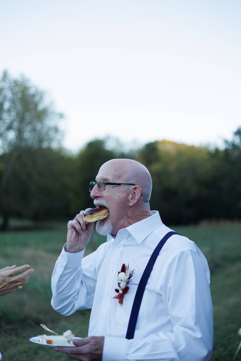 Groom enjoys a donut at an outdoor wedding reception with a plate of cake