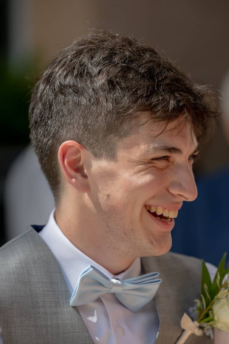 Groom in a grey vest and light blue bowtie laughs joyfully during a wedding ceremony