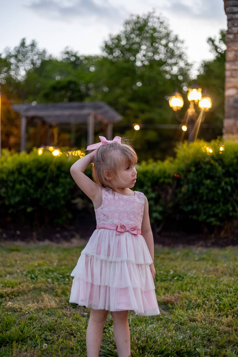 Young girl in a pink dress poses outdoors at dusk with string lights in the background