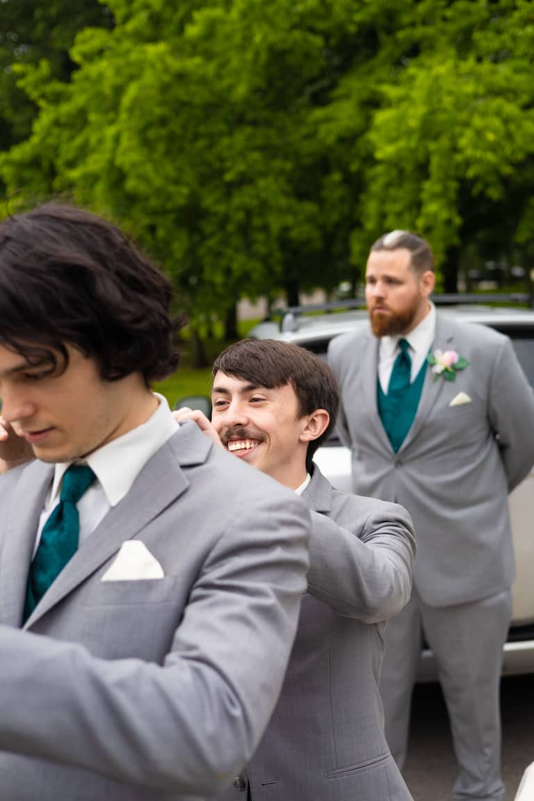 Groomsmen in grey suits with teal ties get ready outdoors on a bright day
