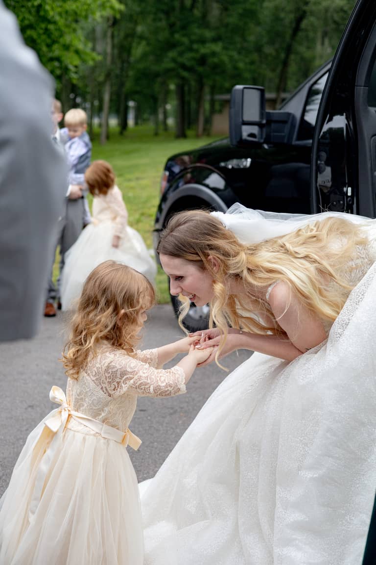 Bride shares a tender moment with a flower girl outdoors by a black truck