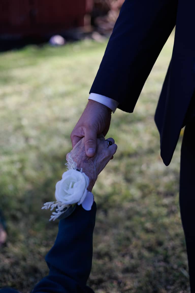 Groom holds grandmother's hand with a white rose corsage at outdoor wedding