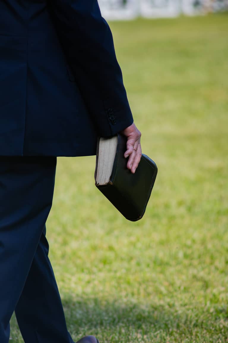 Groom in a dark suit carries a bible across a grassy lawn during a wedding ceremony