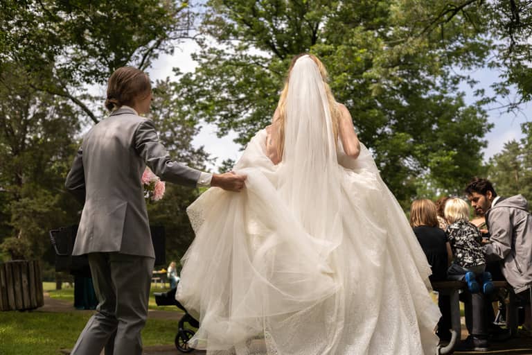 Groom holds bride's train at outdoor wedding with guests watching