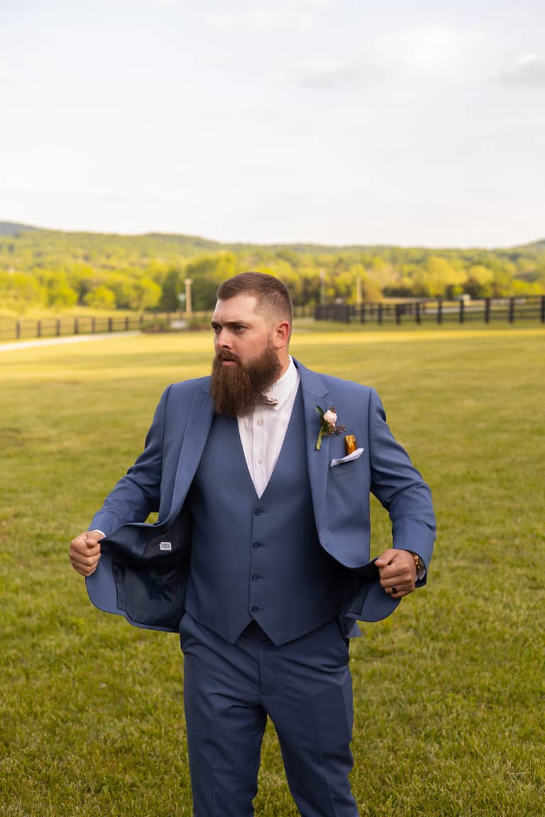 Groom in blue suit adjusts jacket on grassy field with trees in background