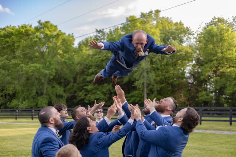Groomsmen in blue suits toss a groom in the air outdoors with trees and a fence behind them