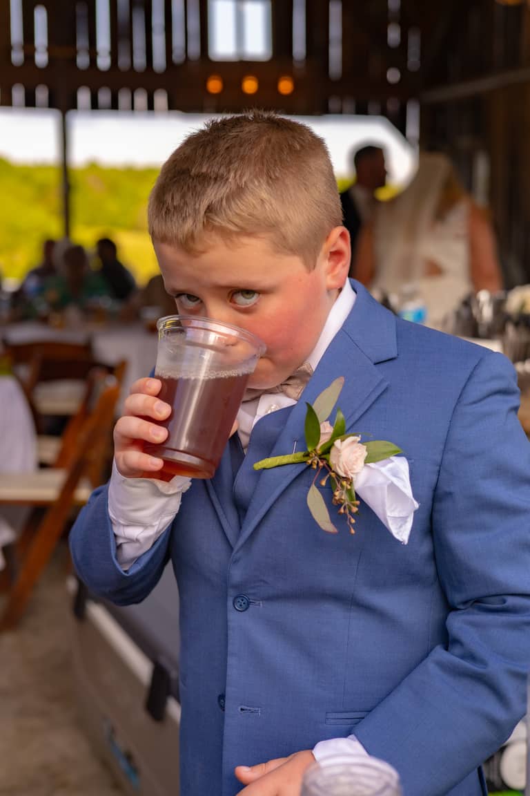 Young ring bearer in a blue suit sips a drink at an outdoor wedding reception