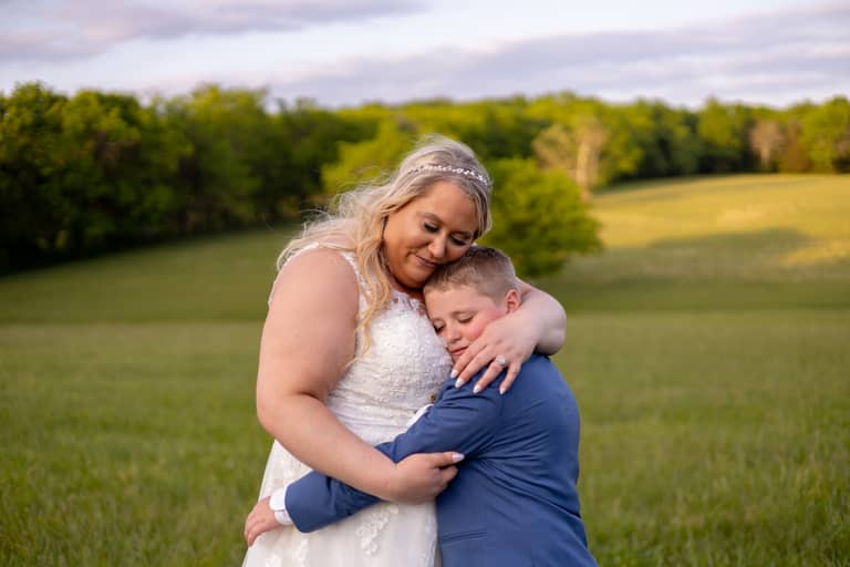 Bride embraces young boy in a tender wedding portrait outdoors at sunset