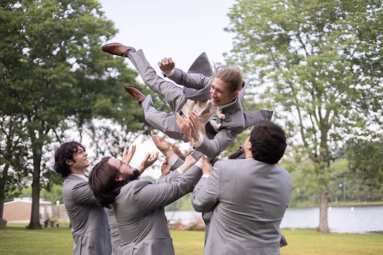 Groomsmen in grey suits joyfully toss the groom in the air outdoors by a lake