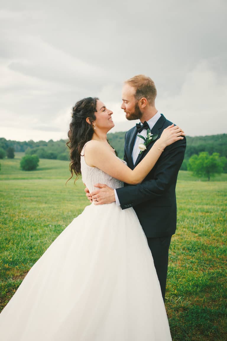 Bride and groom embrace in a grassy field under a cloudy sky, wedding portrait