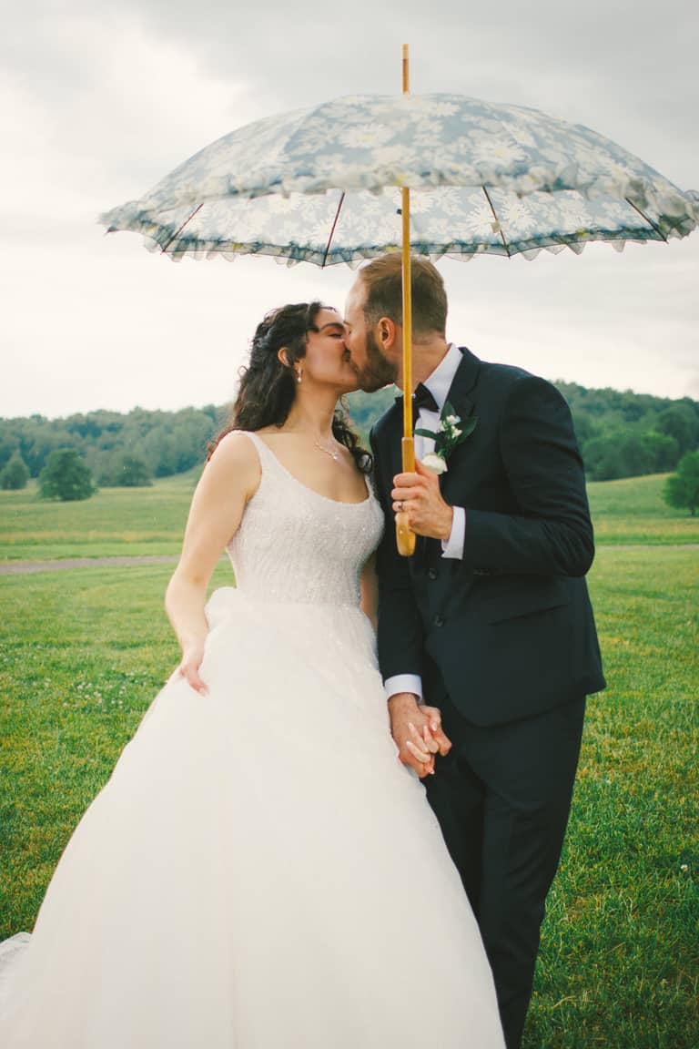 Bride and groom kiss under a floral umbrella on a grassy field during their wedding