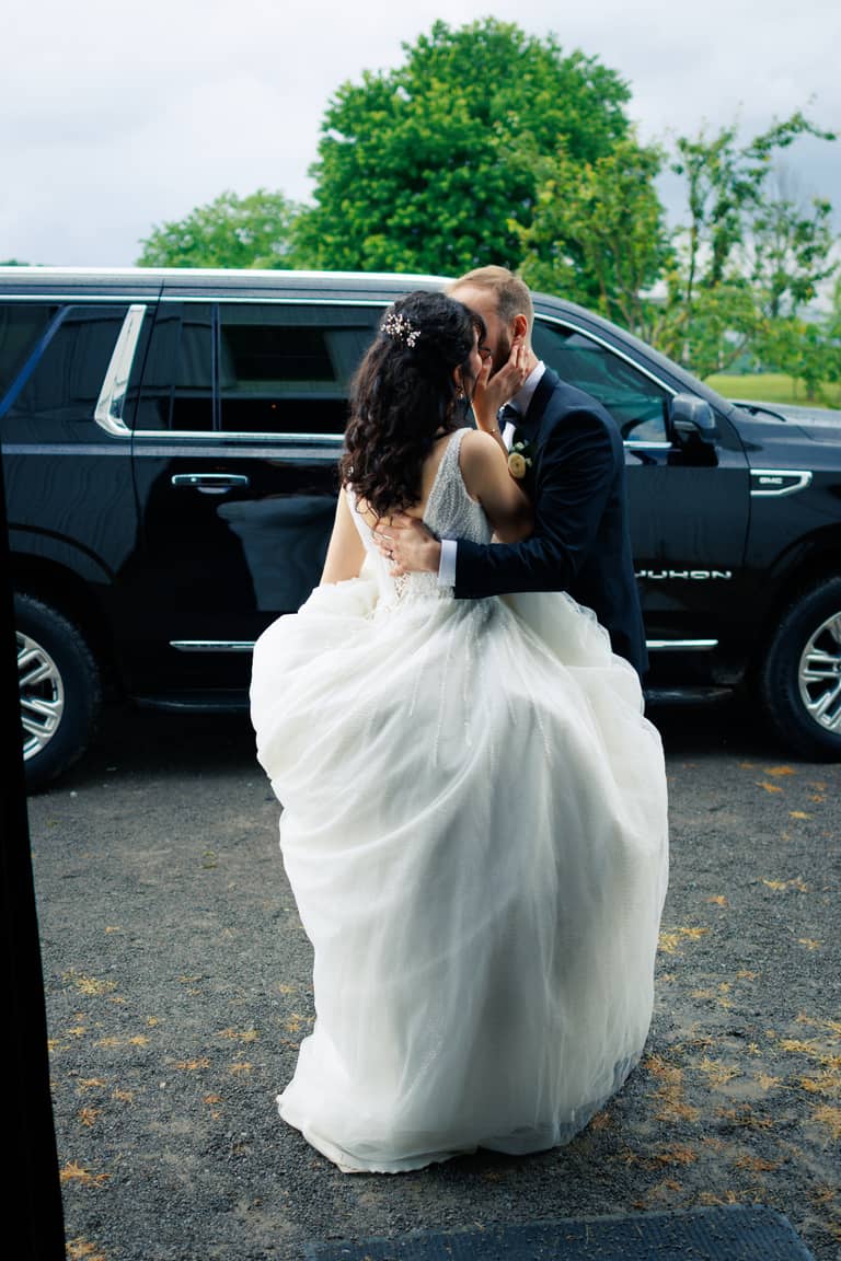 Bride and groom embracing affectionately in front of a black SUV on a cloudy day