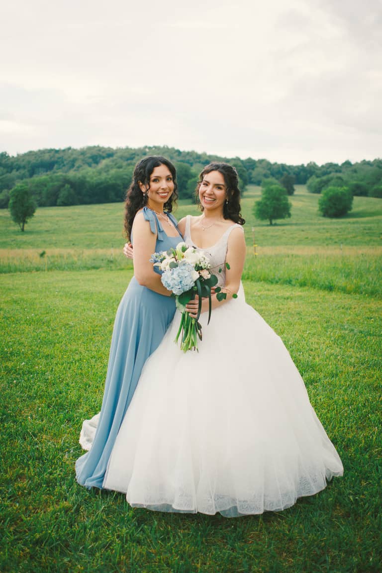 Bride and bridesmaid smile, holding a bouquet in a grassy field wedding portrait