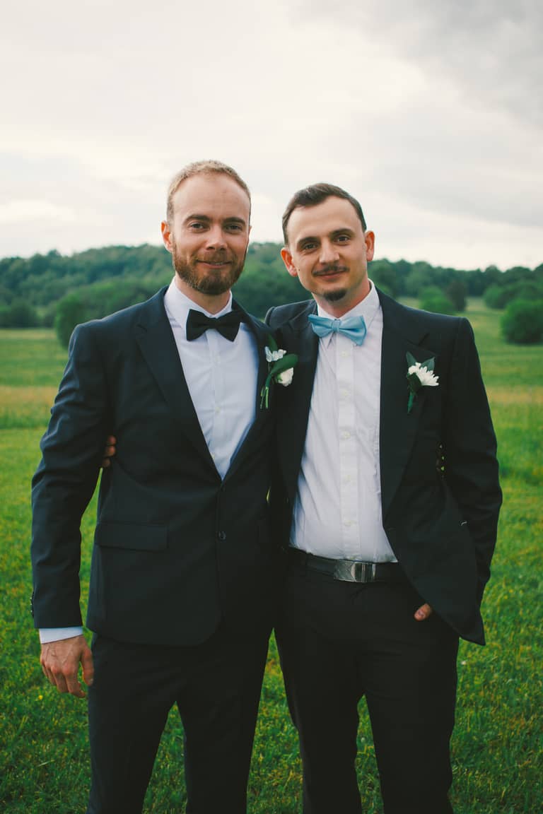 Two grooms in suits smile outdoors with a green, hilly landscape behind them