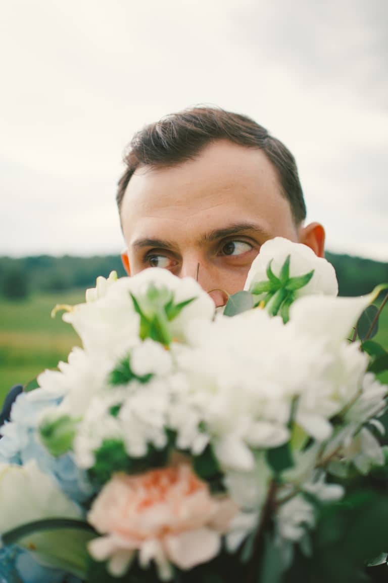 Groom peeking over a bouquet of white flowers on a wedding day