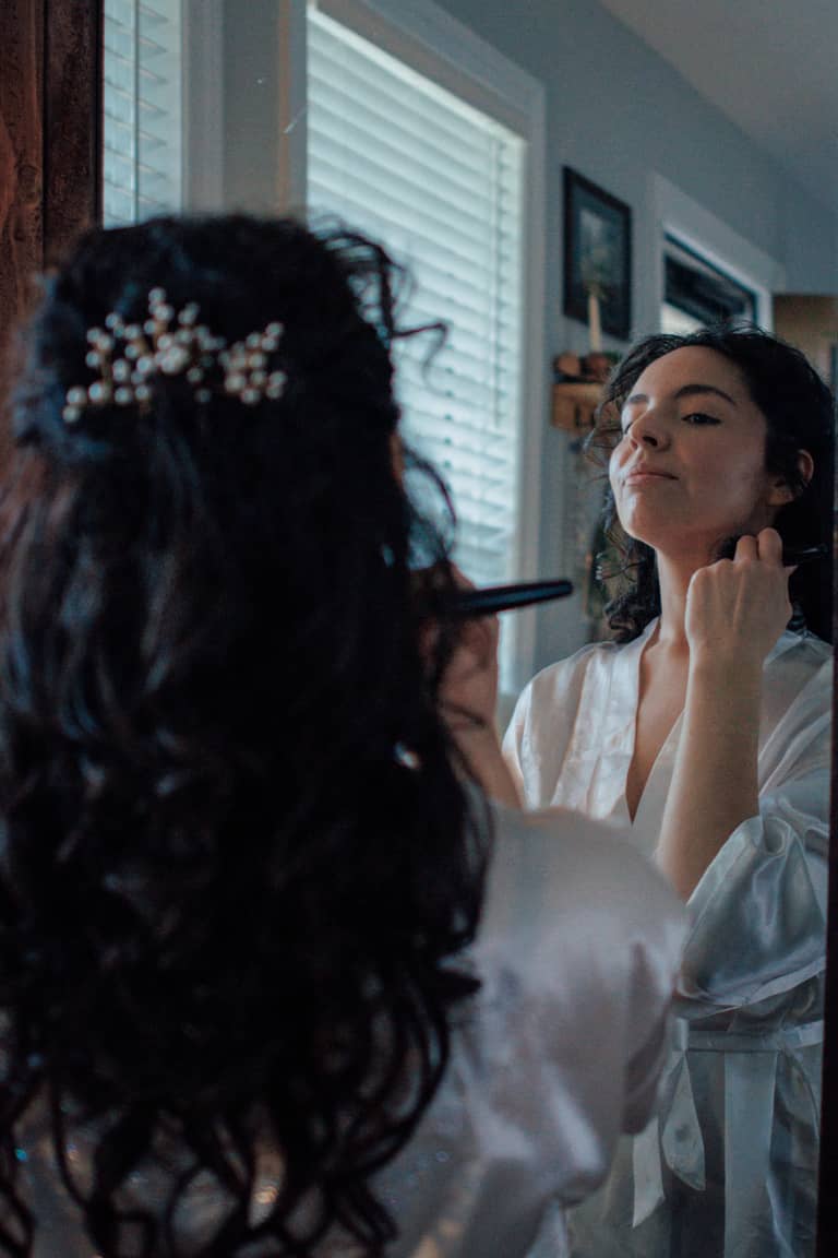 Bride applying makeup in a satin robe, reflected in a mirror, wedding preparation